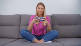 Cheerful gamer girl playing mobile games on smartphone. Happy blonde female sitting on couch and using mobile phone. Beautiful Ukrainian blondie woman plays mobile app game on smart phone