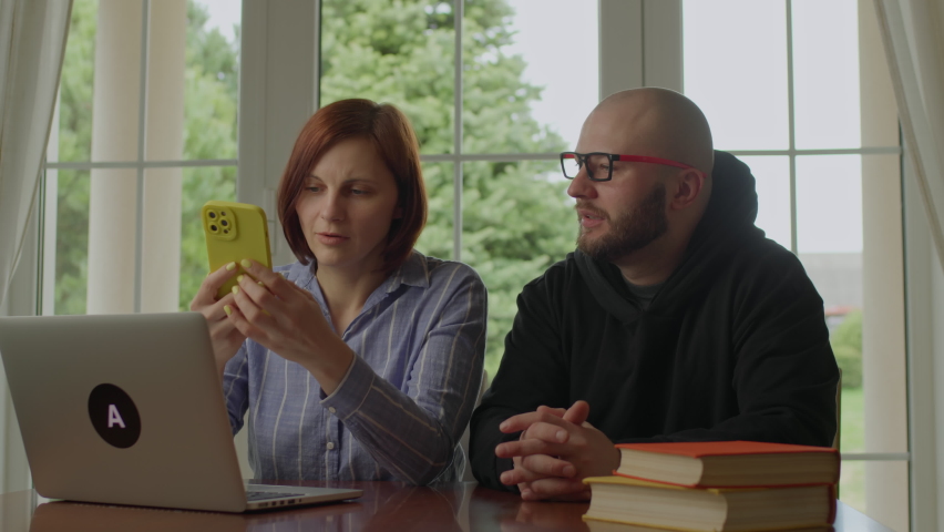 Young adult family spending time together at home. Husband reading paper book and wife using mobile phone.