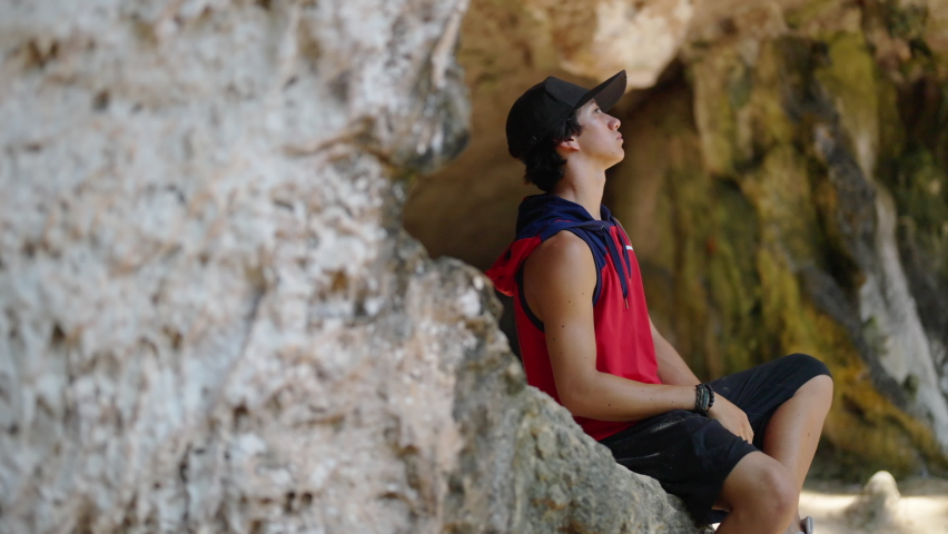Young Asian man solo travel in rock mountain cave on tropical island beach lagoon in summer sunny day. Happy guy relax and enjoy outdoor activity lifestyle on summer holiday vacation trip in Thailand