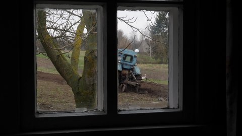Rural view from the window. Old tractor plows a garden, a stork walks around the field and looks for food in the freshly plowed land. Preparation of a summer cottage for planting vegetables, potatoes