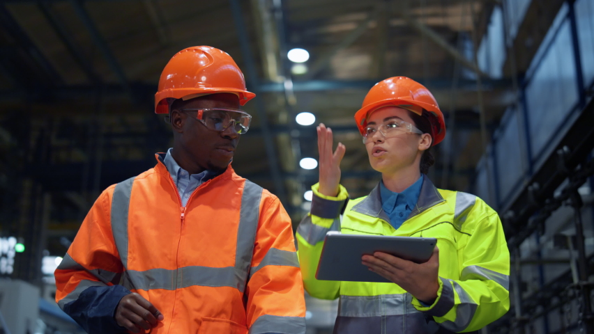 Workers partners holding tablet computer walking at modern manufacturing factory. African american man industrial specialist discussing project work with woman engineer warehouse. Team work concept.