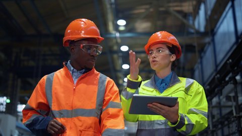 Workers partners holding tablet computer walking at modern manufacturing factory. African american man industrial specialist discussing project work with woman engineer warehouse. Team work concept.の動画素材