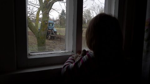 Child girl looks through the window of a rural house at the work of a tractor. Plowing a garden plot with an old tractor. Preparing a garden for planting vegetables, potatoes or cereals. Ukraine