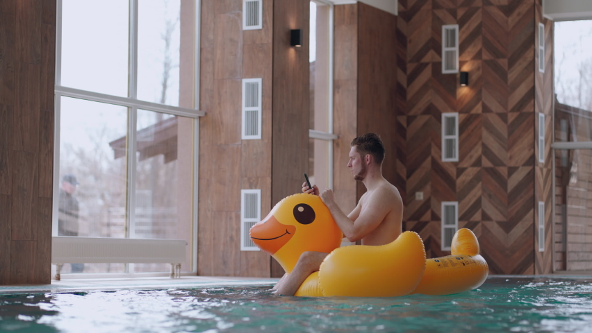 fun and rest in water park, adult man is sitting on inflatable duck in swimming pool and using smartphone