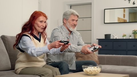 Cheerful senior couple 50-60 years old playing a video game at home sitting on the couch. Game On, Family Meeting, Multi Ethnic Family, Different Generations.