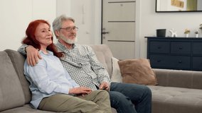 Happy senior couple aged 60-70 watching news on TV while sitting on the couch at home and talking.