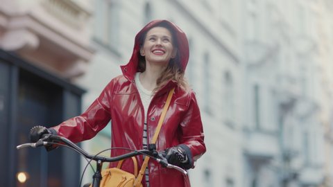 smiling modern 40 years old woman in red rain coat with bicycle in the rain outdoors in the city.