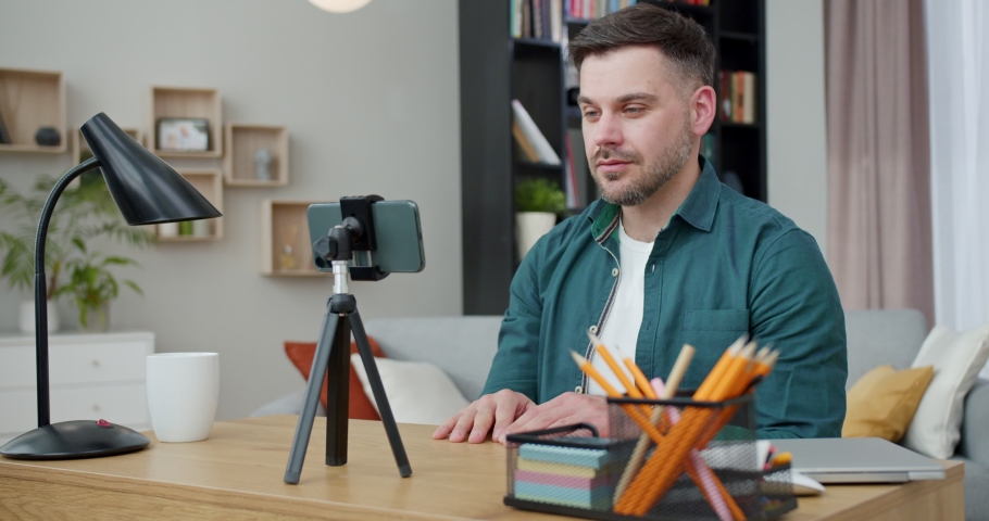 Confident caucasian man sitting at desk and recording video blog on modern smartphone that fixed on tripod. Creation of content for social networks. Royalty-Free Stock Footage #1089569405