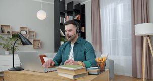 Busy caucasian man in headset sitting at home office with wireless laptop and having video chat. Male freelancer in casual wear taking notes during online meeting.