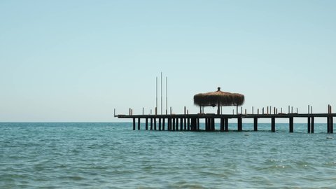 Wooden pontoon in sea, sun rays reflecting in clear sea water surface. Wooden pier above sea water surface with straw umbrellas near luxury hotel complex. Summer beach vacation concept