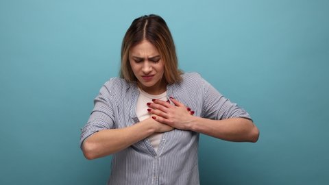 Portrait of unhealthy woman holding hands on chest feeling acute pain in chest, risk of stroke, heart attack, wearing striped shirt. Indoor studio shot isolated on blue background.