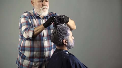 Bearded man coloring hair. Hairdresser is dying male hair. Man dyeing hair while using a brush. Colouring of gray hair at barber salon. Haircare and men hairstyling.