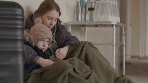 Slowmo of upset young Caucasian refugee woman and her little son sitting close to each other in shelter wearing old warm clothes, having nowhere to go