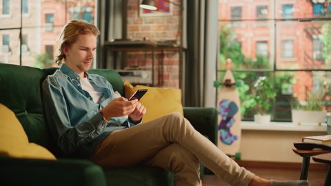 Young Handsome Adult Man Sitting on a Couch in Living Room, Relaxing and Using Smartphone. Creative Male Checking Social Media, Chatting with Friends, Browsing Internet. City View from Big Window.