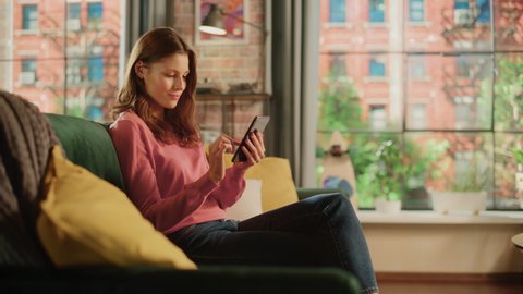 Young Beautiful Female Sitting on a Couch in Living Room, Relaxing and Using Smartphone. Creative Girl Checking Social Media, Chatting with Friends, Browsing Internet. City View from Big Window.