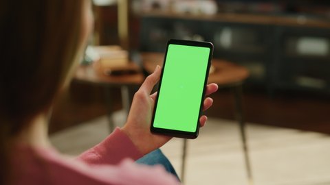 Feminine Hand Scrolling Feed on Smartphone with Green Screen Mock Up Display. Female is Resting at Home and Checking Social Media on Mobile Device. Close Up Over the Shoulder Footage.