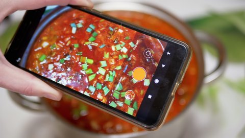 Translation: video clip, video, photo, square. Female hands are recording a video of traditional Ukrainian red borscht on a smartphone. Photographing food with a mobile phone camera. Fingerprints.