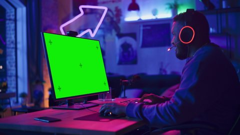 Excited Gamer Playing Online Video Game with a Mock Up Green Screen on His Powerful Personal Computer. Room and PC have Colorful Neon Led Lights. Cozy Evening at Home in Loft Apartment.