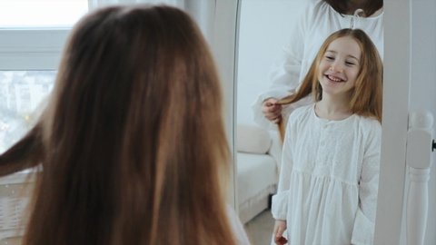 Young mother and cute 6 years daughter are spending time together at home in the morning. Mom combs her little child girl in front of mirror, 4k video