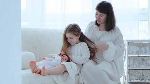 Young adult mother and little cute daughter are spending time together on the sofa at cozy domestic atmosphere. Mom braids hair her child, while she plays with a doll