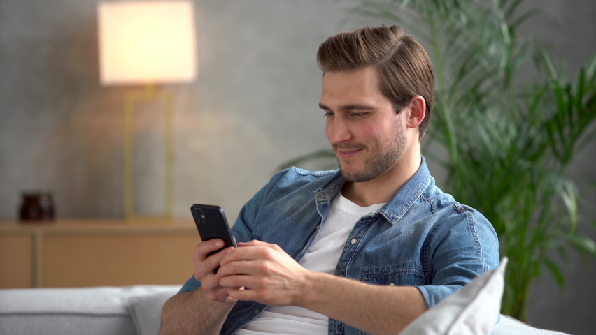Happy Handsome Caucasian Man Using Smartphone in Cozy Living Room at Home. Man Resting on Comfortable Sofa