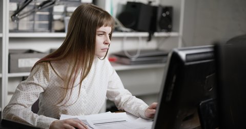 A young woman is sitting at the computer in the office. She looks at the computer monitor then sorts through documents and struggles with sleep. She looks tired and wants to sleep. 