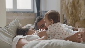 Happy Caucasian couple lying in bed on Sunday morning and playing with their cute baby girl. Adorable infant kissing her mum and dad while parents smiling looking at child. Family, parenthood concept - Powered by Shutterstock - Get 15% off with code: PIKWIZARD15
