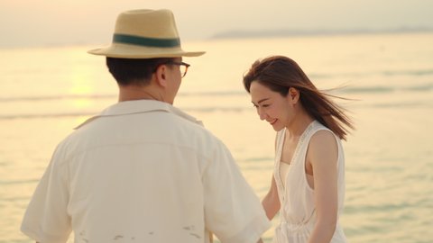 4K Asian couple holding hands and embracing each other while walking on tropical beach together at summer sunset. Happy family husband and wife enjoy outdoor lifestyle on holiday travel beach vacation