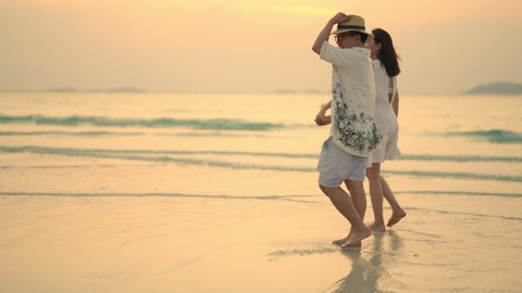 Happy Asian family couple holding hand and walking together on tropical beach at summer sunset on beach vacation. Cheerful husband and wife relax and enjoy outdoor lifestyle on holiday travel vacation