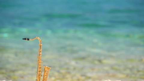Mini model of real alt tenor saxophone stands white small pebbles on seashore, against background of blue turquoise water. Music screensaver for romance. Slow motion video. Copy space for your text.