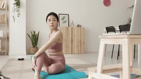 Happy young asian chinese woman sitting on a yoga mat following an online fitness class on laptop - Technology, sports and healthy lifestyle concept.