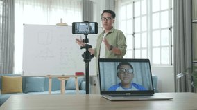 Asian Male Teacher With Glasses Shoots Video By Smartphone Camera While Having Video Call On Laptop And Teaching Math At Home

