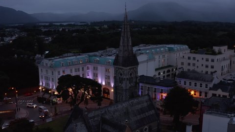 Pull back shot of historic stone church with tall spire. Backwards fly above evening town. Killarney, Ireland