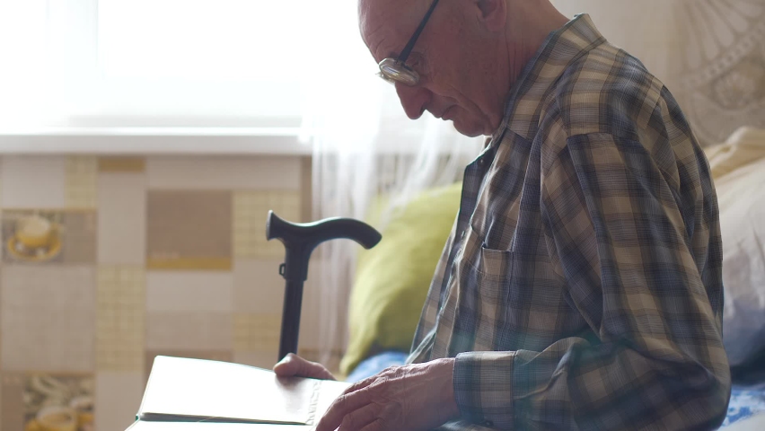 Caucasian Pensioner 70-80 years old with a walking stick at home sitting looking at photos in a family photo album. Nostalgic memories of a pensioner. Family tree in photo album.Selective focus Royalty-Free Stock Footage #1089599947