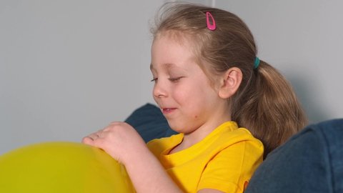 A beautiful child in a yellow t-shirt holds a yellow inflated balloon and blows air into his face. The balloon is deflating. The girl laughs. Children's laughter and joy.
