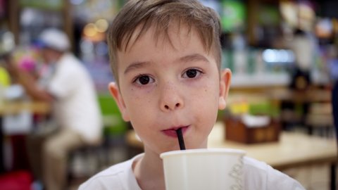 Little boy child against the background of visitors in a cafe looks at the camera and drinks juice. Feeding a kid in a cafe. Son boy on sitting at the table drinking carbonated juice