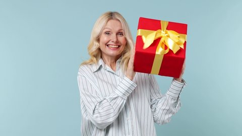 Cheerful elderly blonde woman lady 60s years old in white shirt hold shake red present box with gift ribbon bow try to guess what inside isolated on plain pastel light blue background studio portrait