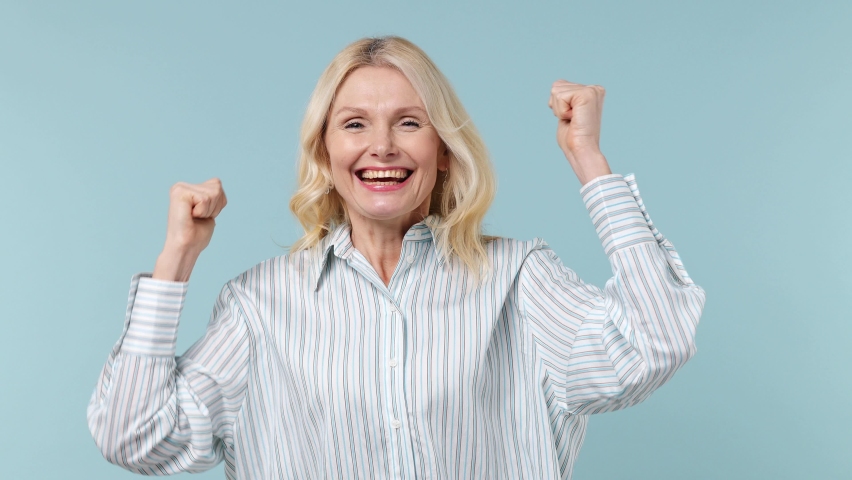 Excited fun elderly gray-haired blonde woman lady 60s years old wears white shirt doing winner gesture celebrate clenching fists say yes isolated on plain pastel light blue background studio portrait