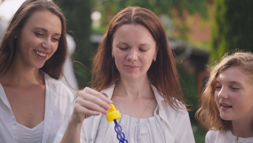 Close-up portrait of joyful pregnant woman blowing soap bubbles with friends sitting outdoors on backyard. Carefree relaxed Caucasian women and teenage girl enjoying leisure in slow motion