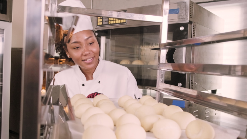 Professional African American female chef in white cook uniform, gloves, and apron making bread from pastry dough, preparing fresh bakery food, baking in oven at restaurant's stainless steel kitchen. Royalty-Free Stock Footage #1089611809