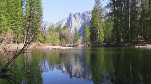 Cathedral Rocks in Yosemite valley reflecting in Merced River with glacier water slowly flowing with rippling surface like mirror. Scenic peaks reflected in clear water surrounded by green pine forest