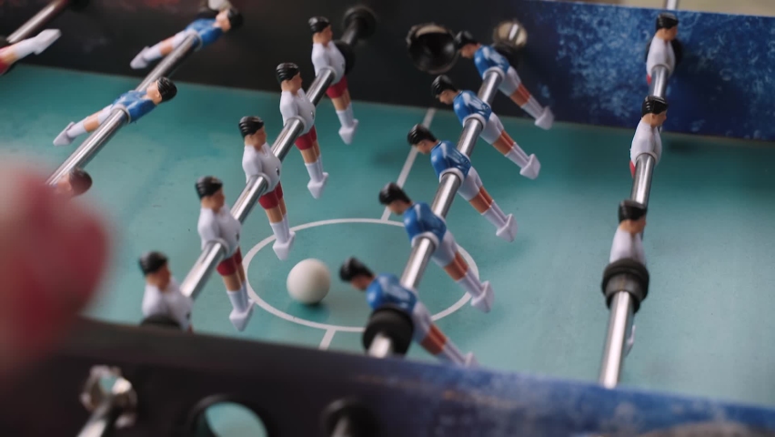 Players introduce white kicker ball into table football game and start playing. They spin metal axles with players and hit ball across field.