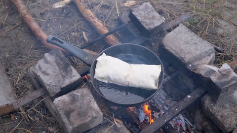 Preparing Meat Shawarma Wrapped in Pita on Outdoor, Burning Bonfire, in Pan. Juicy lavash fries in blazing, smoking flame, fire on a skillet, in woods. Concept of food, hiking, survival in nature.