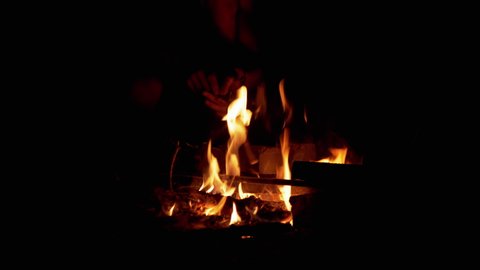 Male Hands, Using of Branch, Kindles a Night Bonfire in the Open Air. Hot fire, smoldering coals, sparks, smoke, ash flying up. Tourist warms himself up by the fire. Concept survival, hiking, trip.