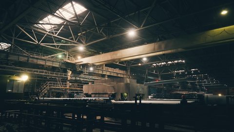 Large girder or beam crane transports iron pipes inside large dark workshop in metallurgical factory