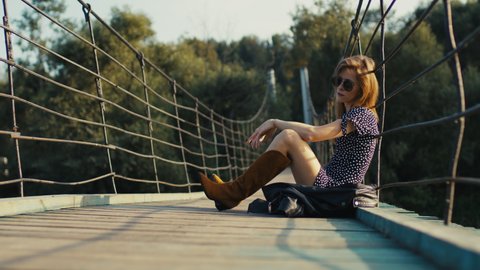 A young, girl in a dress and cowboy boots sits on a suspension bridge at sunset.