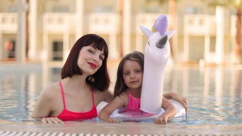Young girl kid with unicorn inflatable ring splashing in swimming pool having fun leisure activity. Happy mother and her daughter in the same pink swimsuits playing in swimming pool. Summer vacations