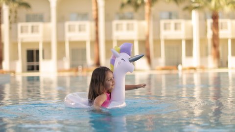 Child in swimming pool waving her hand . Little girl playing in water. Vacation and traveling with kids. Children play outdoors in summer. Kid with unicorn inflatable ring toy.