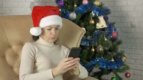 Sad female with phone on Christmas party. A sad female in Santa hat relax and chatting in loneliness in the room with winter decorations.