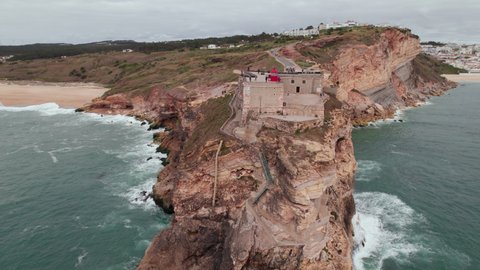 Aerial view of an old lighthouse on a cliff with a fortress on the coast of the Atlantic ocean in Nazare town, Portugal. Drone footage